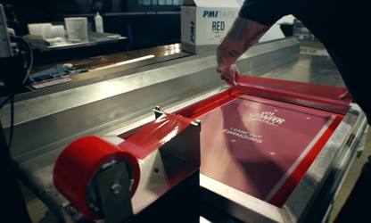 Close-up of a person applying PMI Red Screen Printers Tape to the edge of a screen printing frame in a workshop, with a roll of red tape in a dispenser in the foreground.